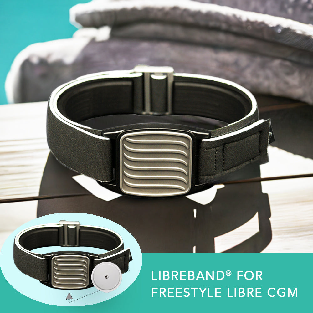 A pewter wave design Freestyle Libre 2 CGM sports and swim armband with a black neoprene strap, displayed on a wooden surface with a reflective shadow below.