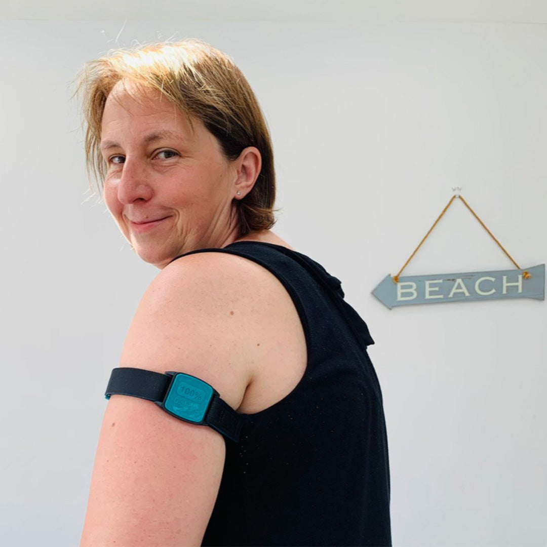 Customer wearing teal Libreband protecting Freestyle Libre sensor on upper arm sitting by sign for beach.